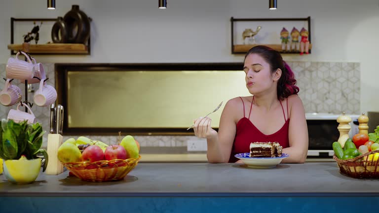 Happy Indian alone smiling girl sitting in modern kitchen eating  piece of cake at indoor house. Overjoyed beautiful cute female celebrate birthday having fun enjoying holiday inside cozy home