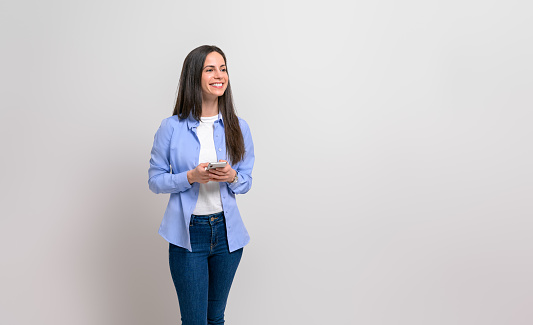 Confident young female entrepreneur messaging over cellphone and looking away on white background