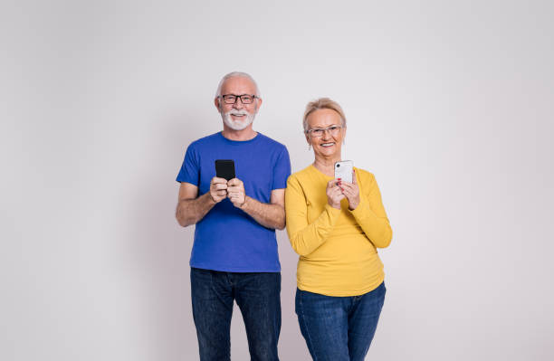 retrato de una pareja mayor sonriente que envía mensajes a través de teléfonos móviles mientras está de pie sobre fondo blanco - senior adult mature adult senior couple heterosexual couple fotografías e imágenes de stock