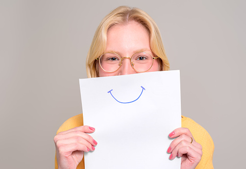 Portrait of young woman in eyeglasses holding white paper with smiley drawing on white background