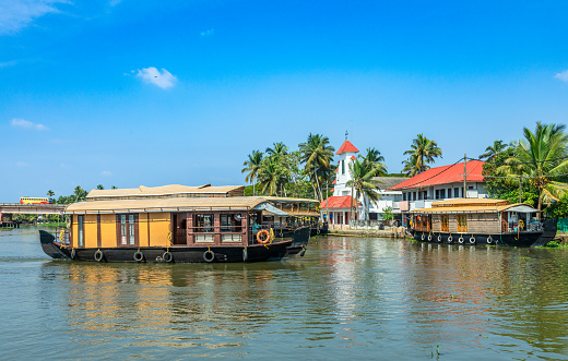 Old colonial Saint Thomas catholic church on the coast of Pamba river, with palms and anchored living houseboats, Alleppey, Kerala, South India