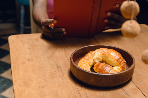 crescents served in deep brown rustic ceramic plate on wooden table with unrecognizable afro woman sitting reading book blurred in background, copy space