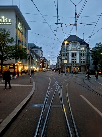 In the Center of the City district Oerlikon in Zurich. The image shows several people walking on a street behind a cable car.