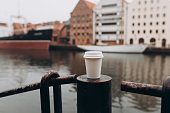 White Paper cup of coffee on bridge near river on city street. Traveling Europe in autumn. Close-up view of disposable paper cup take away, for mockup