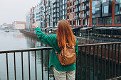 Cheerful redhead woman posing on city street in Gdansk. 30 girl with backpack pointing finger on urban background. Traveling Europe in autumn. Urban lifestyle concept.