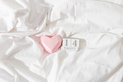 Valentine's Day concept, pink paper valentine heart and 14 february holiday date on wooden calendar in bed on on  white crumpled sheets. Minimal style flat lay photo, top view, copyspace, pastel color