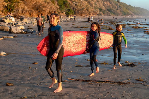 Three girls carrying a long surfboard on the sunset beach. Swami's Beach, Encinitas, Cardiff-by-the-Sea, San Diego area, California. December 28, 2023