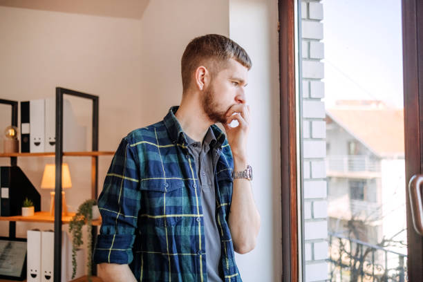istock Young disappointed man looking through window at home 1894687878