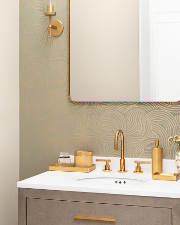 A bathroom sink detail with a gold faucet and decorations, a patterned wallpaper, white marble countertop, and a snake skin cabinet.