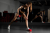 Young fit slim woman does hands muscles exercises with red dumbbells in gym against mirror background