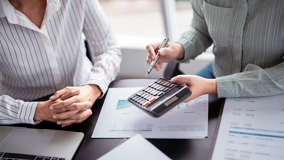 Two business real estate seller calculating about property investment and showing statistic on calculator while explaining terms data document about home sales contract and home insurance in office.