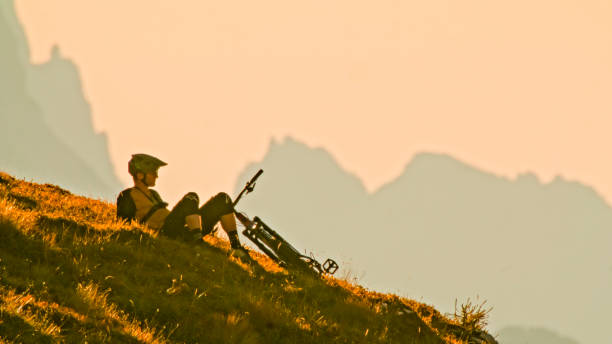 In a Serene Moment,a Thoughtful Male Mountain Biker Relaxes on a Hill against the Backdrop of a Clear Sky During the Mesmerizing Hues of Sunset. The Scene Captures the Tranquility of Nature and the Contemplative Pause of an Adventurer