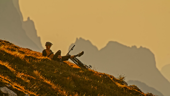 In a Contemplative Moment,a Thoughtful Male Mountain Biker Relaxes with His Foot Up on the Cycle,Perched on a Hill against the Canvas of a Clear Sky During the Enchanting Hues of Sunset. The Scene Captures the Serene Connection Between the Rider and Nature