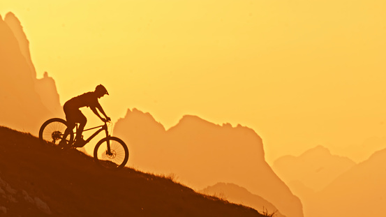 Graceful Ride at Sunset,a Side View of a Silhouette Male Athlete Riding his Mountain Bike on a Hill against the Majestic Backdrop of Mountains and a Clear Sky During the Enchanting Colors of Sunset. The Scene Embodies the Blend of Athleticism and the Tranquil Beauty of Nature
