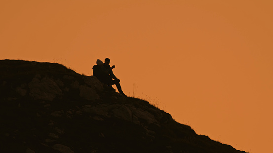 Silhouetted against a Clear Orange Sky During the Tranquil Sunset,a Hiking Couple Finds Serenity Sitting on a Mountain Peak. The Scene Encapsulates the Shared Joy and Peace Discovered amidst the Beauty of Nature's Twilight Embrace