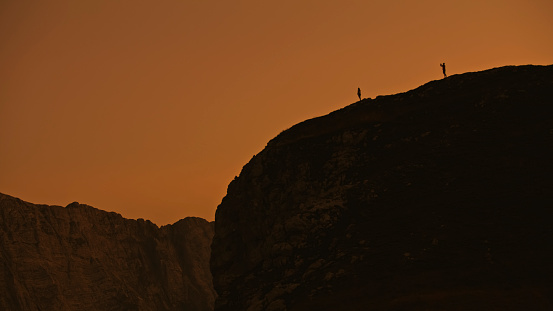 In a Low Angle,Distant Silhouette,Climbers Stand atop a Colossal Rocky Mountain against the Backdrop of a Clear Sky During the Mesmerizing Sunset. The Scene Radiates the Sense of Triumph and Awe Inspiring Accomplishment in the Face of Nature's Grandeur
