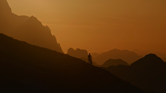 In a Captivating Mid Distance Scene,the Silhouette of a Cyclist Stands on a Hill,Framed against Idyllic Foggy Mountain Ranges and an Orange Sky. This Enchanting Tableau Captures the Cyclist as a Solitary Figure,Surrounded by the Serene Beauty of Nature and the Warm Hues of the Setting Sun