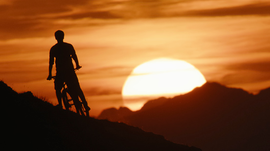 Against an Idyllic Sun and a Dramatic Orange Sky,a Silhouette Male Mountain Biker Stands with his Bicycle on a Hill. The Scene Captures the Essence of Serenity and Triumph,Blending the Thrill of Adventure with the Tranquil Beauty of the Natural Surroundings