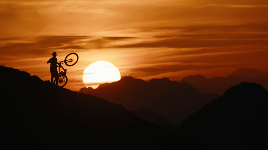 Against a Dramatic Orange Sky,a Silhouette of a Male Mountain Biker is Captured From the Rear,Carrying his Bicycle on a Hill. The Powerful Image Conveys the Determination and Resilience of the Rider against the Striking Backdrop of the Evening Sky