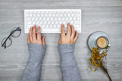 top view of desktop, close up of female hands typing on keyboard, cup of coffee and gypsophila flowers with eyeglasses