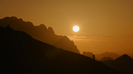 In the Distance,a Silhouette Biker Stands on a Valley Floor against the Backdrop of Majestic Mountain Ranges and an Orange Sky During Sunset. The Image Portrays the Solitary Figure in Awe-Inspiring Surroundings