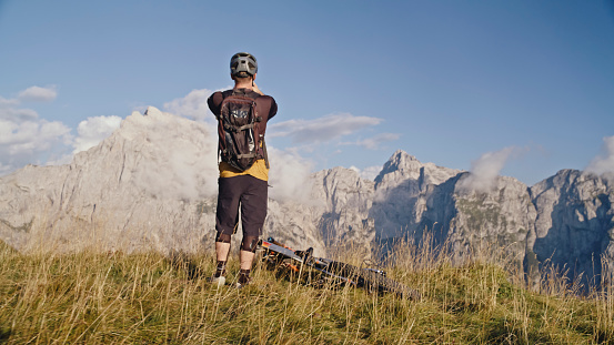 In a Rear View,a Male Biker Stands by his Cycle on a Grassy Field against the Backdrop of Rocky Mountains during a Sunny Day. This Picturesque Scene Captures the Essence of Outdoor Adventure and the Beauty of the Natural Landscape