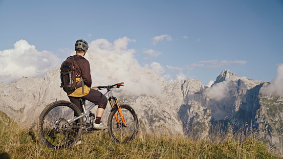 In a Captivating Rear View,a Male Biker Sits on his Bicycle against the Stunning Backdrop of Rocky Mountains during a Sunny Day. This Scene Captures the Tranquility and Adventurous Spirit amidst the Breathtaking Natural Landscape