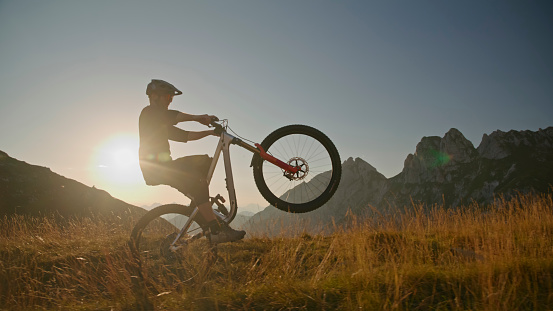 In a Dynamic Side View,Witness the Determination of a Mountain Biker as he Rears Up his Bicycle on a Grassy Field against the Canvas of a Bright Blue Sky during the Mesmerizing Hues of Sunset. This Scene Captures the Intense Skill and Adventurous Spirit in the Magical Twilight