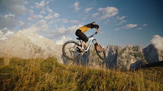 In a Determined Side View,a Skilled Biker Fearlessly Performs a Stunt with his Bicycle on a Grassy Field against the Backdrop of Rocky Mountains and the Expansive Sky. This Scene Encapsulates the Daring Spirit and Precision of Extreme Biking