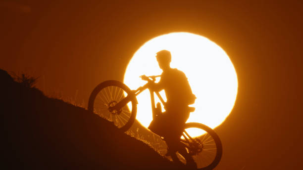 Ascending Silhouette,a Determined Mountain Biker Pushes Bicycle Uphill against the Majestic Sun in a Clear Evening Sky during Sunset. This Powerful Image Conveys the Challenging Spirit and Dedication of the Ascent