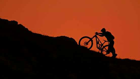Silhouetted against a Clear Orange Sky,a Determined Mountain Biker Pushes his Bicycle while Ascending a hill amidst Majestic Mountains. This Scene Encapsulates the Perseverance and Adventure of Conquering the Heights against the Vibrant Canvas of a Sunset Lit Sky