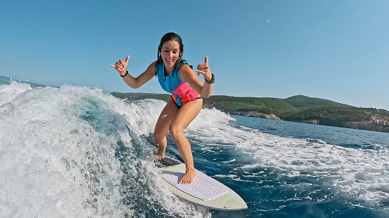 Woman Gesturing Horn Sign while Wakeboarding on the Sea,Her Joyful Expression Capturing the Essence of an Exciting Day with Enjoyable Watersport Adventure,Surrounded by the Refreshing Waves and the Beauty of Nature.