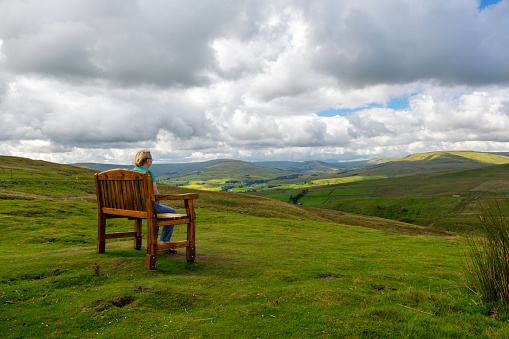 Rear view of a senior woman sat on a bench at a viewpoint in the Yorkshire Dales National Park, England.