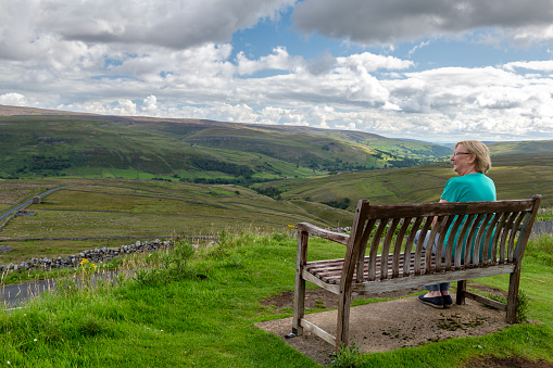 Rear view of a senior woman sat on a bench at a viewpoint in the Yorkshire Dales National Park, England.