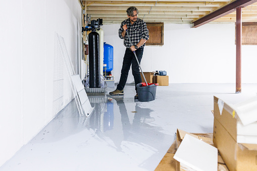 Cleaning the floor after basement flooding. Mature man in plaid shirt removing water after basement flooding. Wet cardboard boxes are on foreground