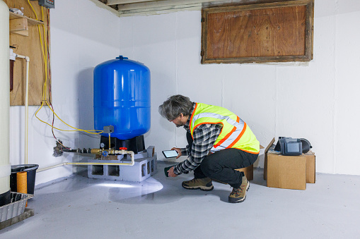 Mature inspector in safety vest, with smartphone and flashlight, examining water pressure tank damage on the flooded floor