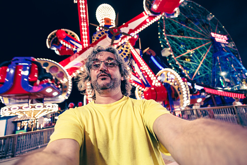 Mature Caucasian bearded man in eyeglasses. grimacing on selfies and having fun at nighttime amusement park
