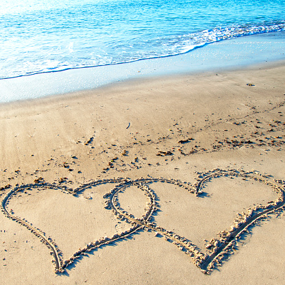 The hand writing heart shaped on the beach by the sea with white waves and blue sky background