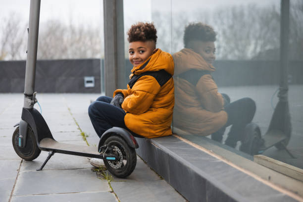 Child in orange takes a moment to reflect alongside their electric scooter, capturing a peaceful and thoughtful scene. Concept of electrical city mobility.