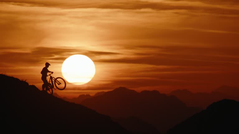 SLO MO Lockdown Shot of Silhouette Mountain Biker Carrying and Pushing Bicycle while Moving Uphill against Dramatic Sunset Sky