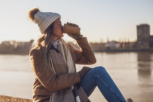 Beautiful woman in warm clothing enjoys  drinking coffee and resting by the river on a sunny winter day.