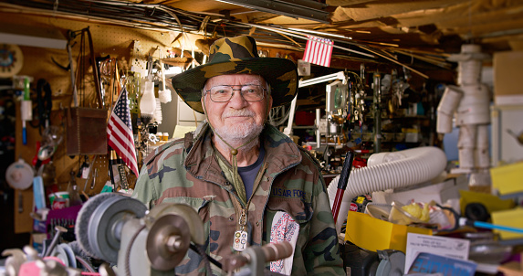 Portrait of an elderly Caucasian man in his garage in Pennsylvania. He is wearing a US Air Force camouflage jacket and a brimmed hat.