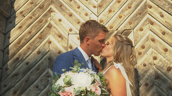 CLOSE UP: Newlyweds in love share a romantic kiss in front of large wooden gates. Beautiful young couple sealed their love with marriage. Romantic looks and tenderness between happy bride and groom.