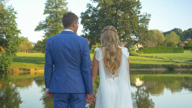 Beautiful couple on their wedding day holding hands while looking at the lake Beautiful couple on their wedding day holding hands while looking at the lake. Authentic romance by the pond with newlyweds. Candid moments full of love between handsome bridegroom and lovely bride. real wife stories stock pictures, royalty-free photos & images