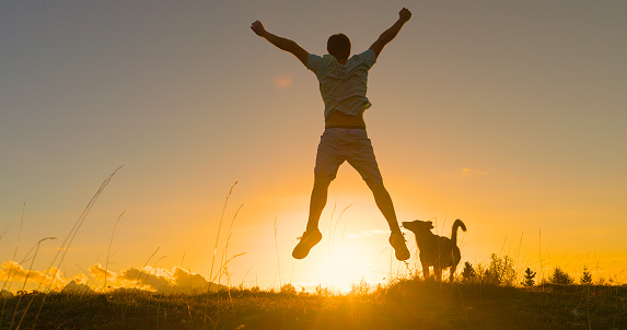 LENS FLARE: Young guy jumps with raised arms on a grassy mountaintop at sunrise. He is happy as he reaches the top with his dog when first rays of autumn sun start spilling over alpine mountains.