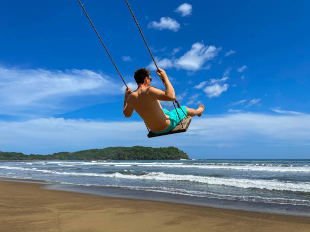 istock LOW ANGLE VIEW Playful young man on a beach swing overlooking wavy Pacific Ocean 1895527883