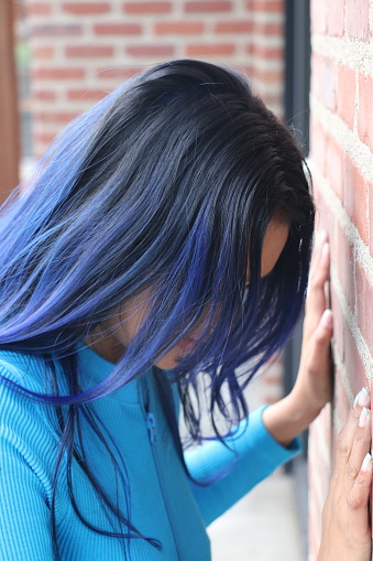 A closeup of a Fijian woman banging her head against a brick wall.  She is wearing long, blue straight hair and a blue tracksuit.