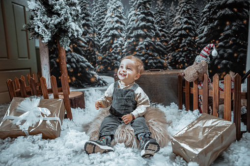 Cute Smiling Male Baby Enjoying Sitting On Fake Snow Among Toys, Presents And Christmas Atmosphere