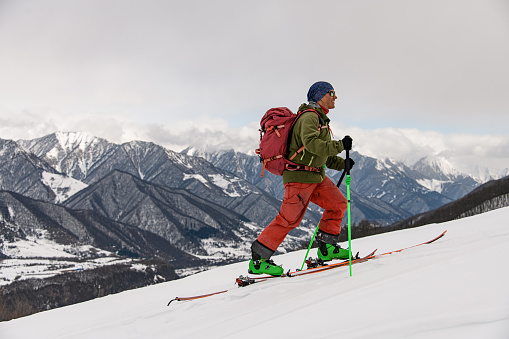 side view of male skier with ski equipment walking at snow against the backdrop of magnificent winter mountain landscape and sky