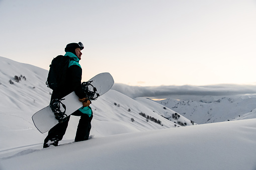 side view of man with snowboard in his hands walks at snow-covered slope against the backdrop of beautiful winter mountain landscape